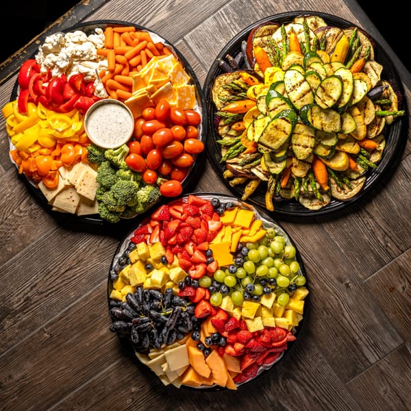 Three large platters of fresh and grilled vegetables, cheeses, and fruits on a wooden table.