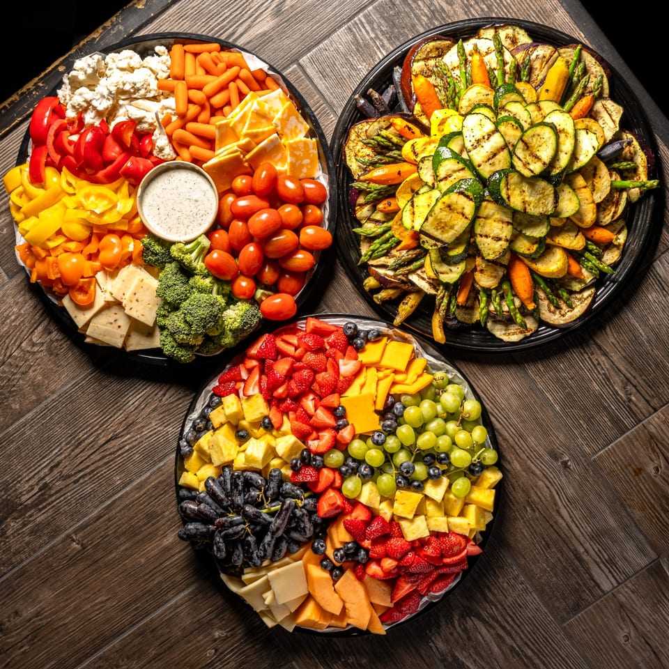 Three large platters of fresh and grilled vegetables, cheeses, and fruits on a wooden table.