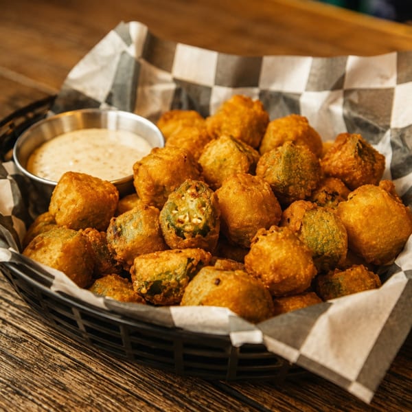 A basket of golden-brown fried okra with a side of creamy dipping sauce on a wooden table.