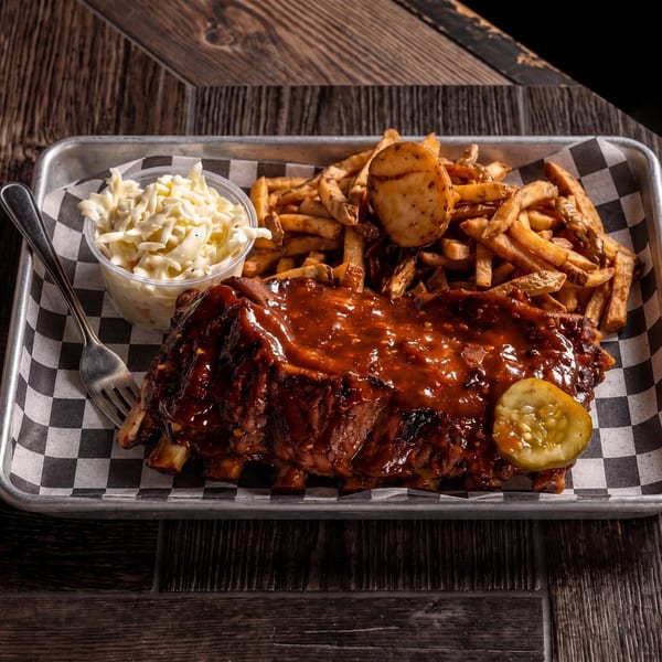 A rack of BBQ ribs, fries, coleslaw, and a pickle slice on a checkered paper-lined tray.